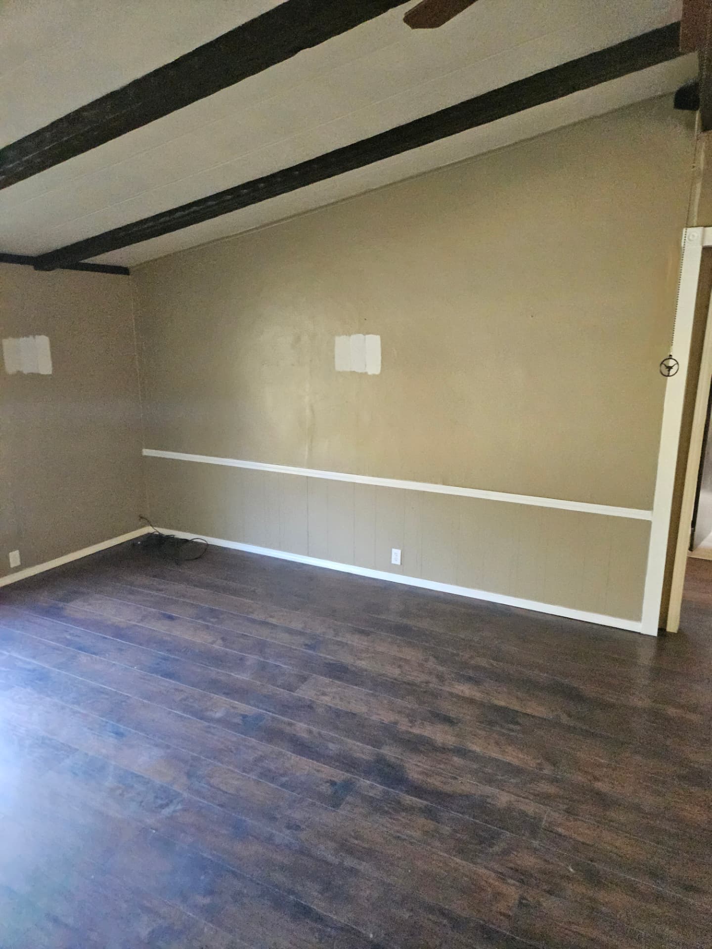 Empty room mid remodel with patched and primed wall spots, tan paneling, white chair rail, and dark ceiling beams before painting in a Johnson County home